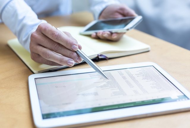 person using a stylus on a tablet with financial data, holding a smartphone in the other hand.