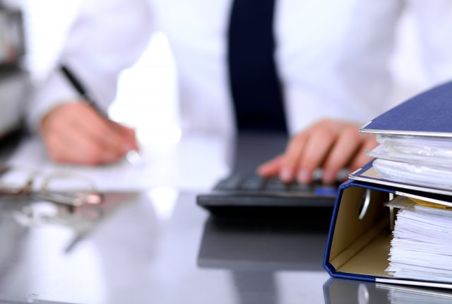 stack of binders filled with documents on a desk, with a businesswoman in the background using a calculator.