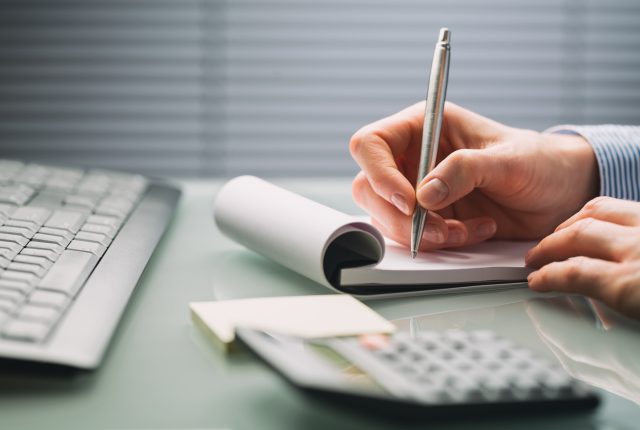 close up of a hand writing in a notepad next to a calculator on a desk.