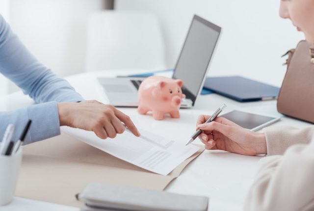 two people discussing paperwork at a desk with a laptop and pink piggy bank in the background