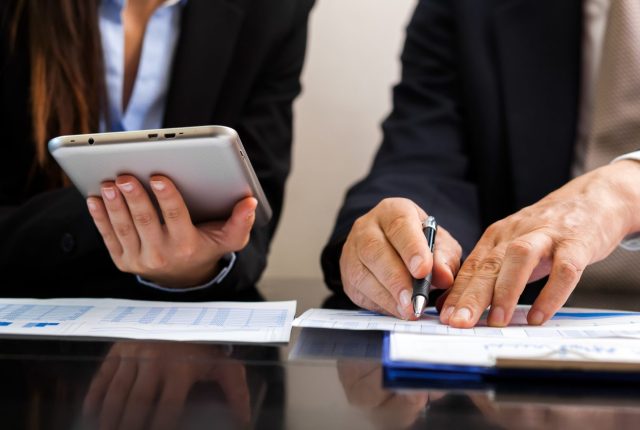 two professionals reviewing documents and using a tablet during a bookkeeping consultation with a small business. They are wearing suits and pointing to figures on a wooden table