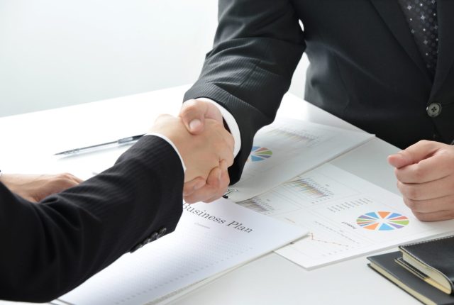 Two business professionals shaking hands over documents with charts and a business plan on the table.
