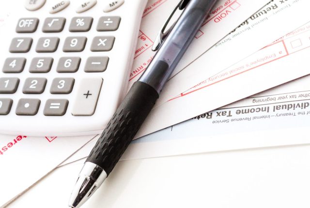 Close-up of a pen, calculator, and eyeglasses resting on tax and income documents.
