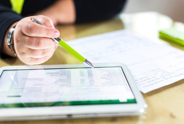 person reviewing financial data on a tablet with a green pen, surrounded by printed spreadsheets on a wooden desk