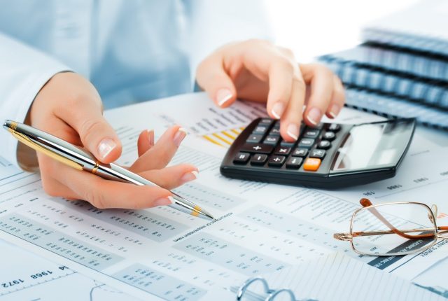 accountant using a calculator while holding a pen over financial documents, with reading glasses and reports on the desk